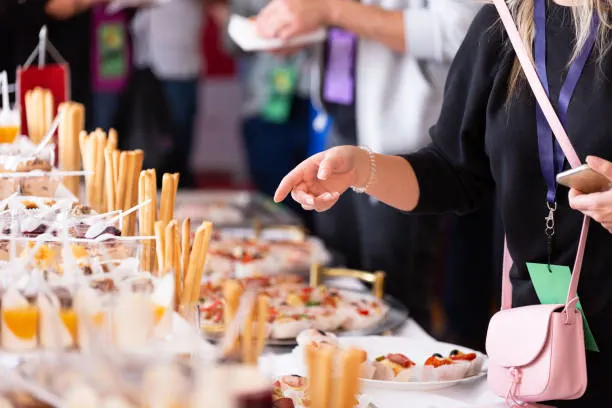 Una persona, vestida con un suéter negro, señala un bufé lleno de aperitivos y postres en un evento. En la mesa hay una gran variedad de opciones, incluyendo palitos de pan, mini pizzas y copas con postres.