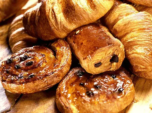  Un primer plano de una variedad de pasteles de panadería, incluyendo croissants y pain au chocolat, apilados sobre una superficie de madera. La iluminación resalta la textura dorada y hojaldrada de los pasteles.