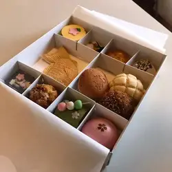 Una caja de cartón blanca abierta muestra un surtido de pequeños dulces y pasteles japoneses, conocidos como wagashi, organizados en compartimentos. Los dulces varían en forma y color, incluyendo un taiyaki (pastel con forma de pez) y un dango de tres colores.