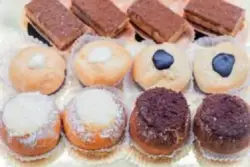  Una bandeja con dos filas de pasteles pequeños. La fila superior contiene pasteles de chocolate con forma de barra, mientras que la fila inferior muestra pasteles redondos cubiertos con glaseado y diferentes toppings, como arándanos y trozos de chocolate.